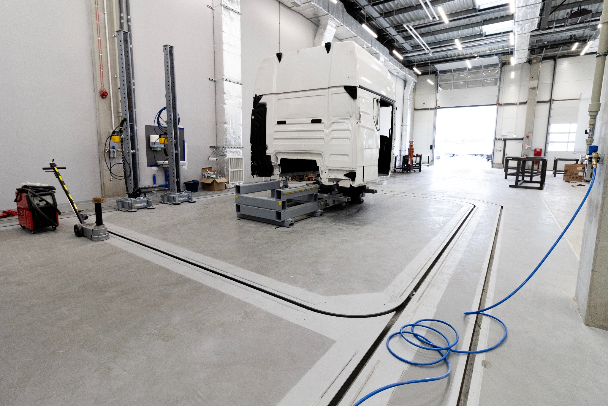 Wnętrze hali warsztatowej z kanałem naprawczym i podnośnikami, przygotowane do naprawy kabiny ciężarówki. Widoczne narzędzia i oświetlenie.