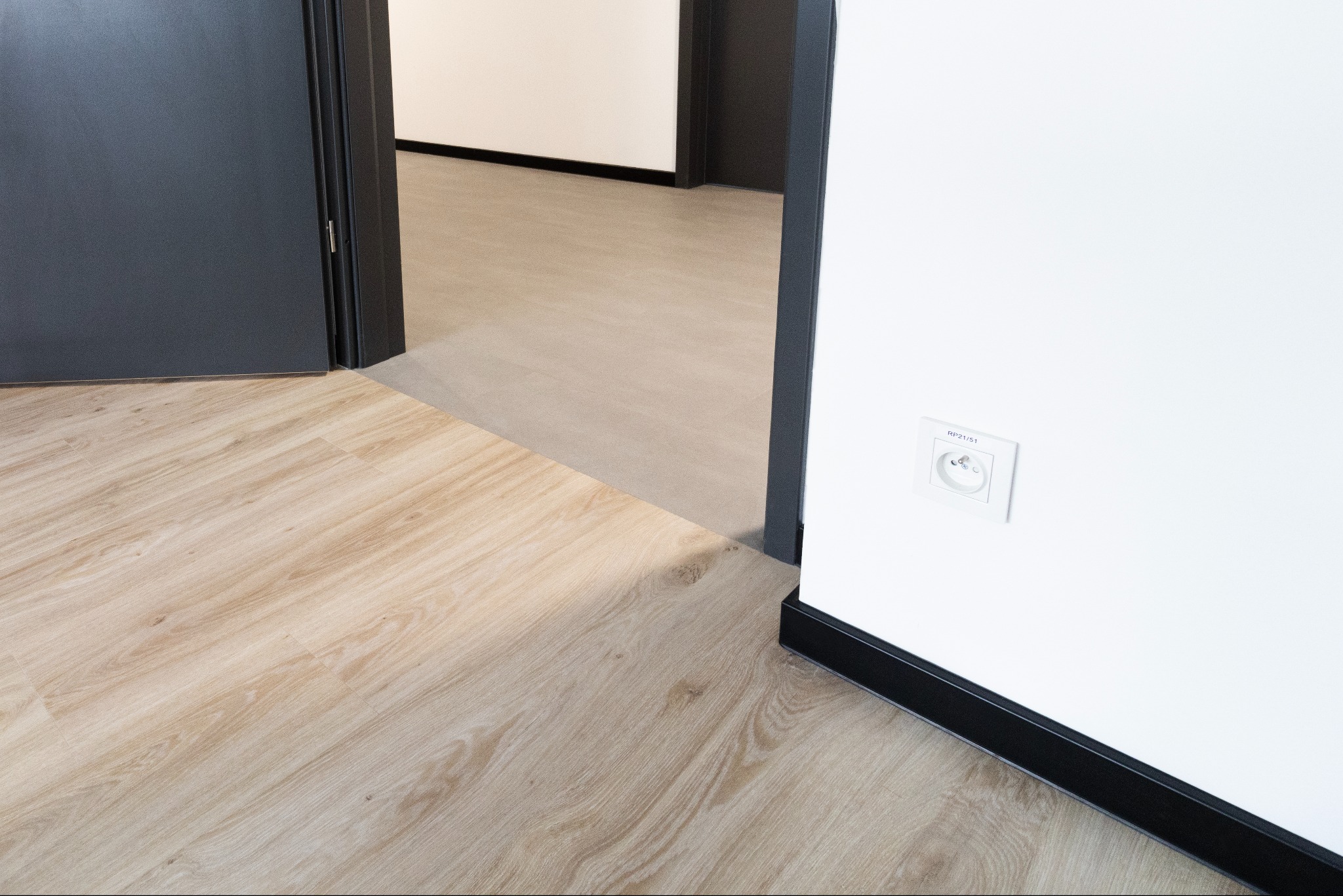 Jasne panele podłogowe w nowym wnętrzu, widoczne przez otwarte, ciemne drzwi. Biała ściana z gniazdkiem elektrycznym, czarna listwa przypodłogowa.