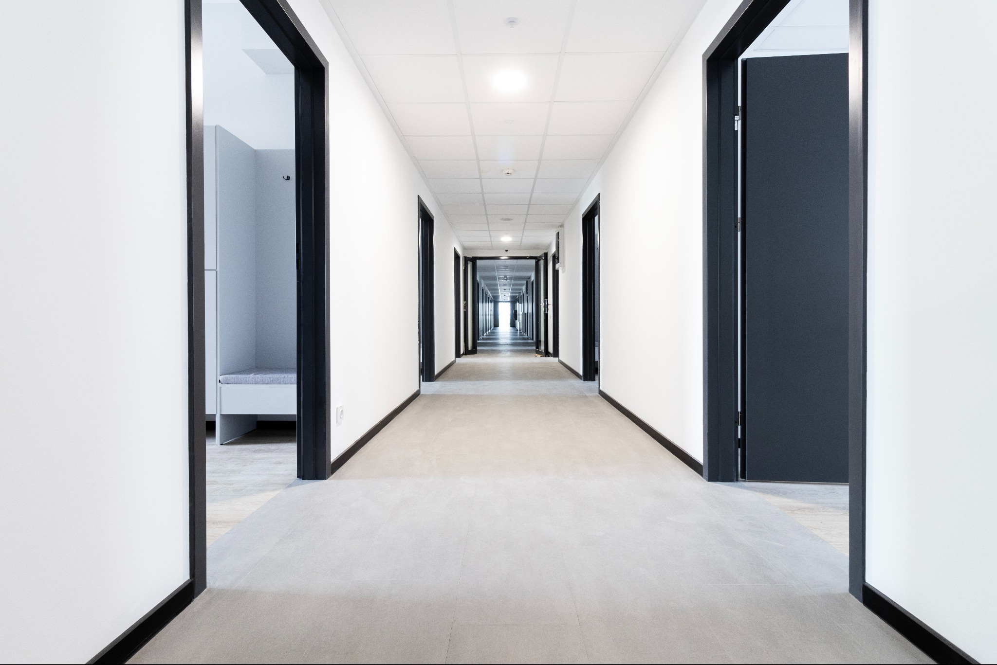 Długi, jasny korytarz z szarą posadzką i czarnymi framugami drzwi, perspektywa zbieżna. Widoczne wejście do pomieszczenia z białą szafką i siedziskiem.