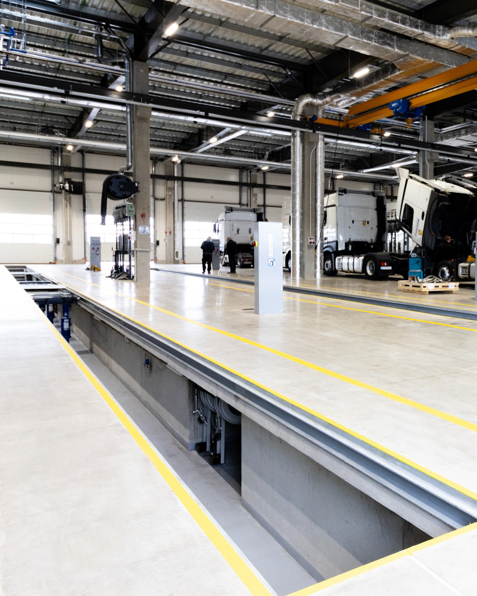 Hala warsztatowa z kanałem naprawczym, betonową posadzką i żółtymi pasami. W tle widoczne naprawiane samochody ciężarowe oraz pracownicy serwisu.