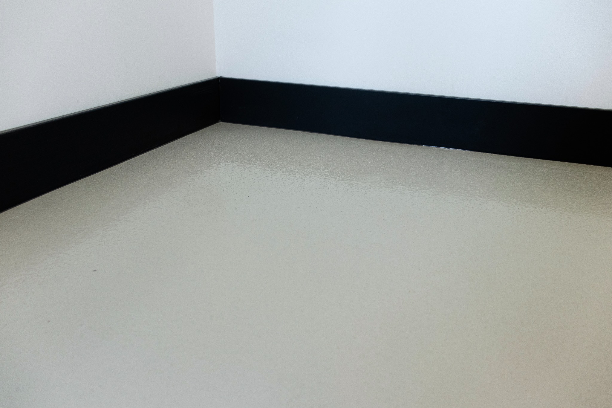 Jasna posadzka żywiczna w rogu pomieszczenia z czarną listwą przypodłogową. Gładka powierzchnia, minimalistyczny design, neutralna kolorystyka.