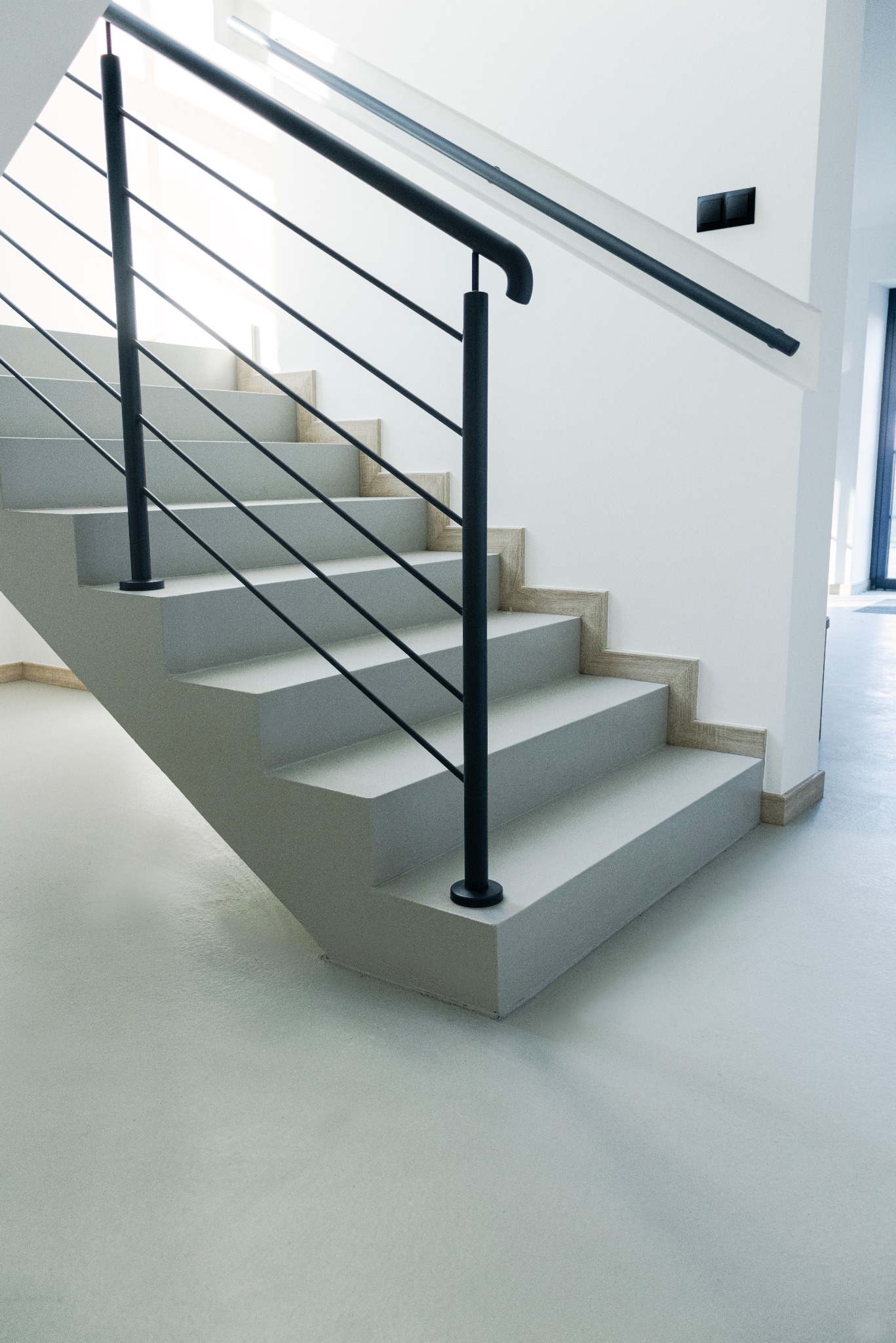Minimalistyczne, jasnoszare schody z czarną balustradą w nowoczesnym wnętrzu. Widoczna gładka posadzka i fragment okna w tle. Stonowana kolorystyka.