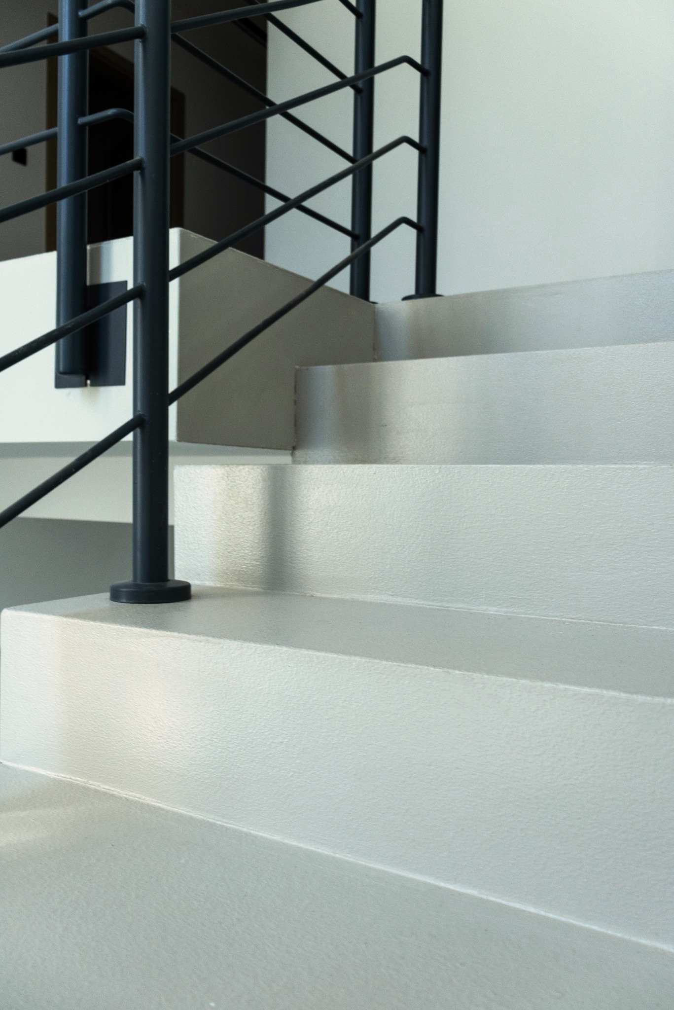 Betonowe schody z czarną, metalową balustradą, oświetlone naturalnym światłem. Widoczne trzy stopnie z delikatną teksturą i minimalistyczny design.