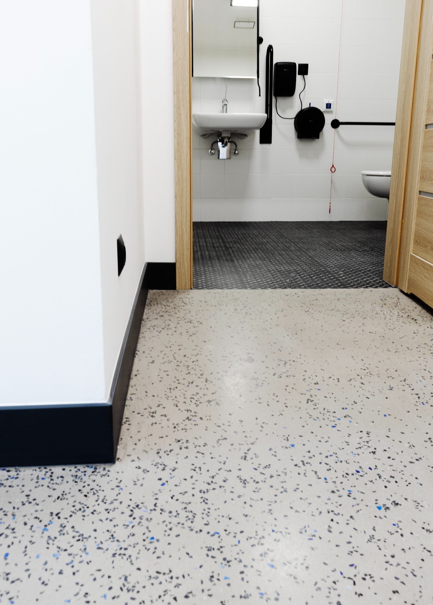 Beżowa posadzka żywiczna z kolorowymi drobinkami w publicznej toalecie z szarą podłogą w tle. Widok na umywalkę i toaletę dla niepełnosprawnych.