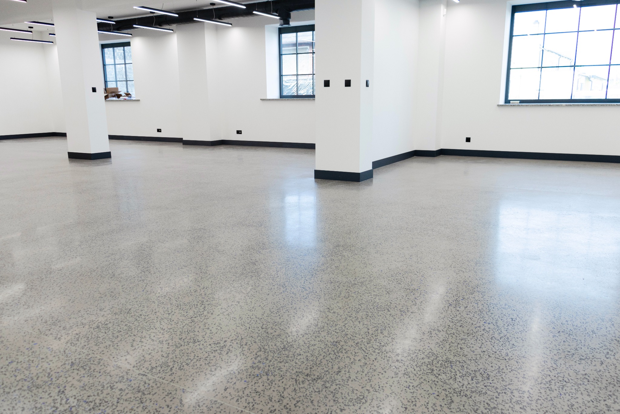 Wnętrze z szarą, polerowaną posadzką z widocznymi ziarnami, białe ściany z czarnymi listwami przypodłogowymi, kolumny i okna z widokiem na zewnątrz.