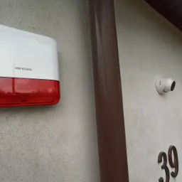 Zewnętrzny alarm dźwiękowy Hikvision z czerwonym światłem i kamera monitoringu zamontowane na szarej ścianie budynku obok brązowej rury spustowej i numeru domu 39.