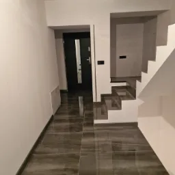 Korytarz z błyszczącą, ciemną podłogą z płytek imitujących kamień, białe ściany i minimalistyczne schody z białymi stopnicami. W tle widoczne czarne drzwi z długim, pionowym przeszkleniem.