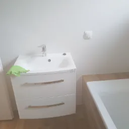 Nowoczesna łazienka po remoncie: biała szafka pod umywalkę z chromowaną baterią, obok wanna z drewnopodobnymi panelami, widoczny włącznik światła na ścianie i zielona ściereczka na szafce.