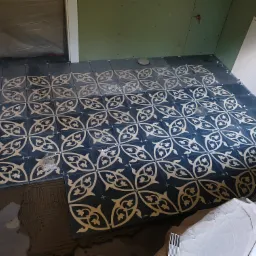 Układanie ciemnych płytek podłogowych z jasnym, dekoracyjnym wzorem w łazience, widoczne krzyżyki dystansowe i świeży klej.