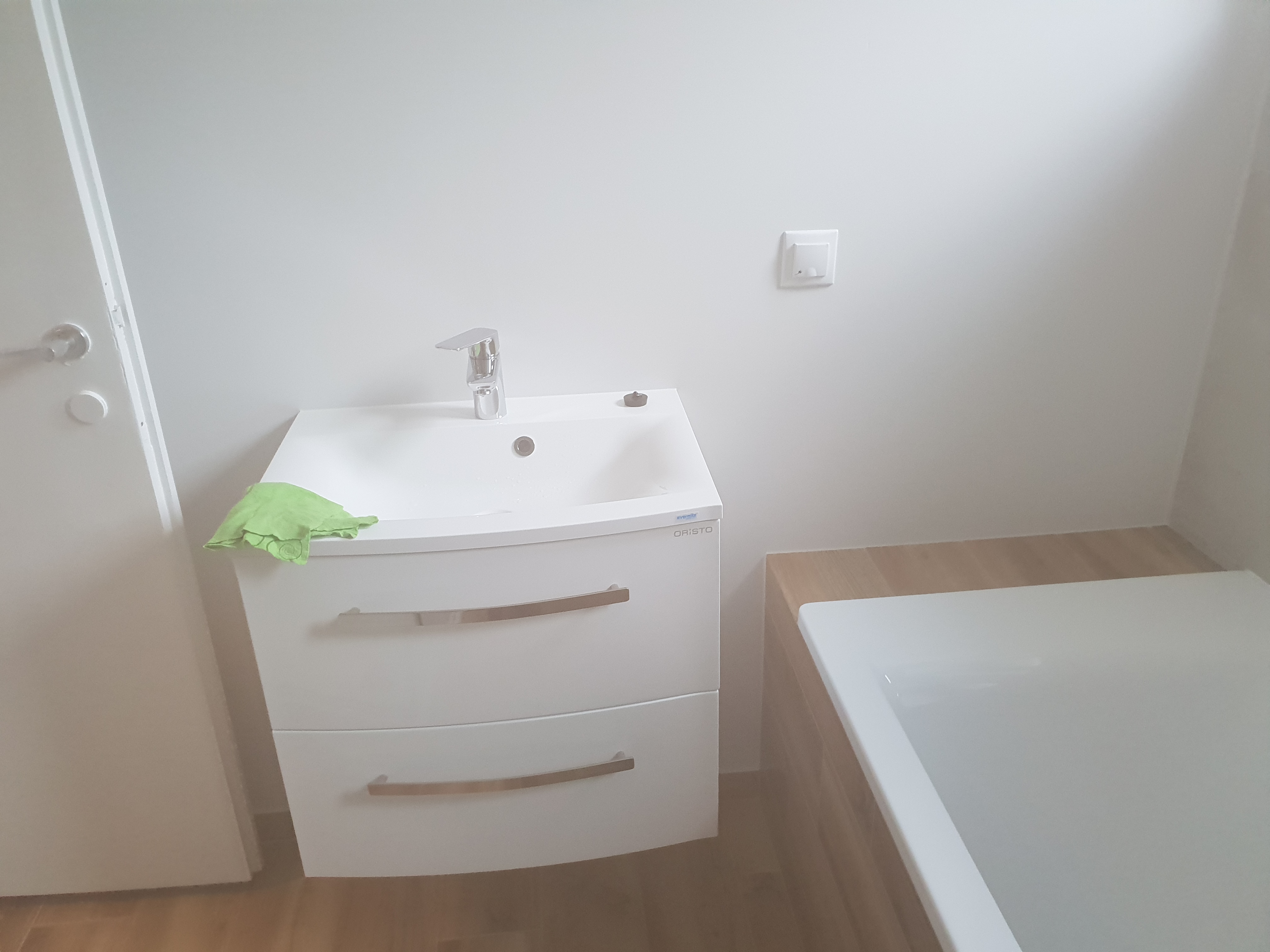 Nowoczesna łazienka po remoncie: biała szafka pod umywalkę z chromowaną baterią, obok wanna z drewnopodobnymi panelami, widoczny włącznik światła na ścianie i zielona ściereczka na szafce.