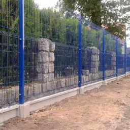 Długi, niebieski system ogrodzeniowy z paneli z drutu, z betonową podmurówką i słupkami obłożonymi kamieniami, zakończony drutem kolczastym, otoczony zielenią.