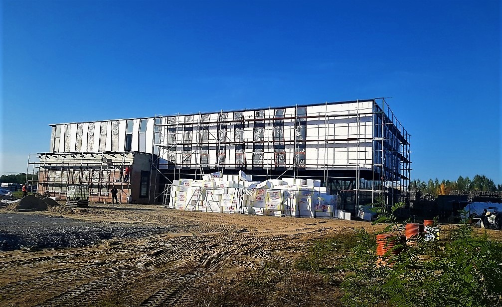 Budynek w trakcie ocieplania, widoczne rusztowania, warstwy izolacyjne i materiały budowlane na placu budowy, błękitne niebo w tle.