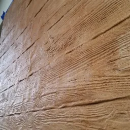 Elewacja domu z fakturą imitującą drewno, widoczna z boku, z fragmentem orynnowania i dachu w tle.