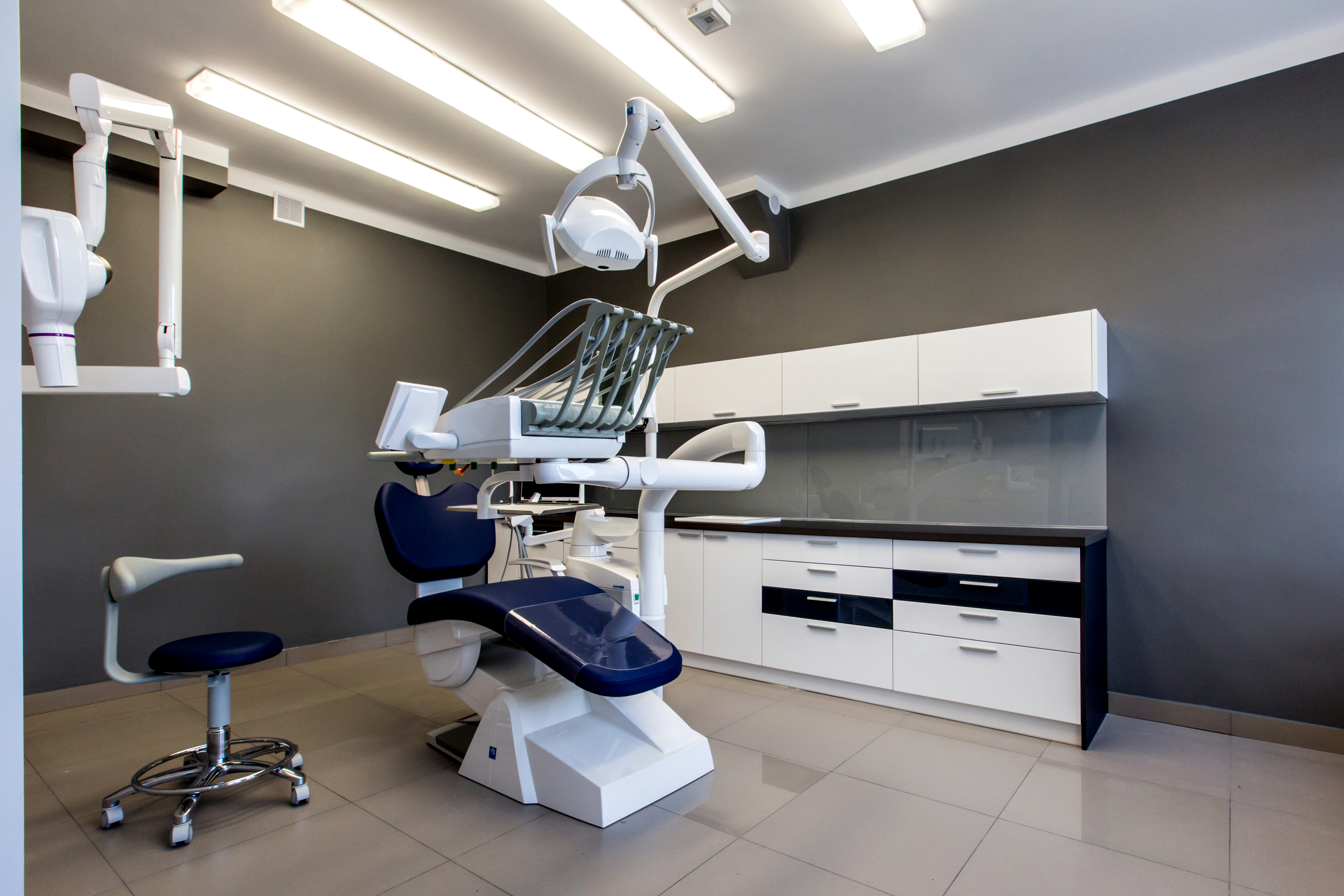 Nowoczesny gabinet stomatologiczny: białe meble na tle szarej ściany, fotel dentystyczny z niebieskim obiciem, oświetlenie sufitowe. Minimalistyczny design, czysta przestrzeń.