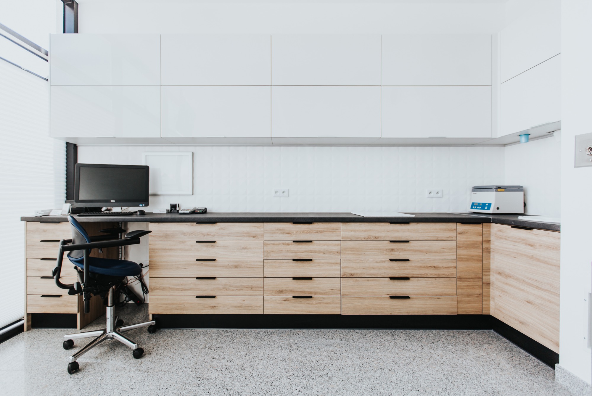 Jasne drewniane szafki z czarnymi uchwytami w biurze, uzupełnione białymi szafkami wiszącymi, biurkiem z komputerem i krzesłem biurowym na kółkach.