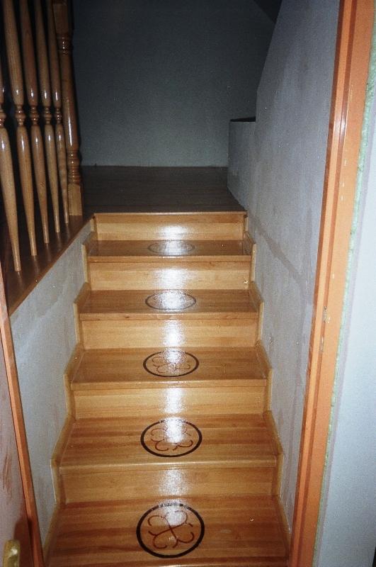 Drewniane schody z dekoracyjnymi motywami czterolistnej koniczyny na stopniach, widoczne z dołu, z balustradą po lewej stronie i ścianą po prawej.