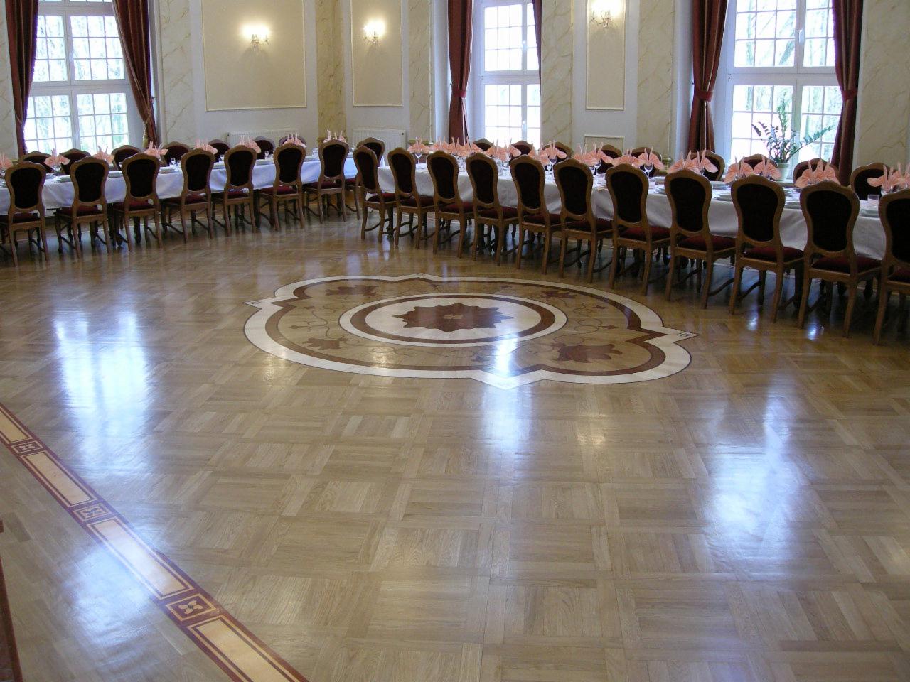 Sala balowa z parkietem w jodełkę, dekoracyjnym wzorem centralnym i długim stołem z krzesłami przygotowanym na uroczystość.