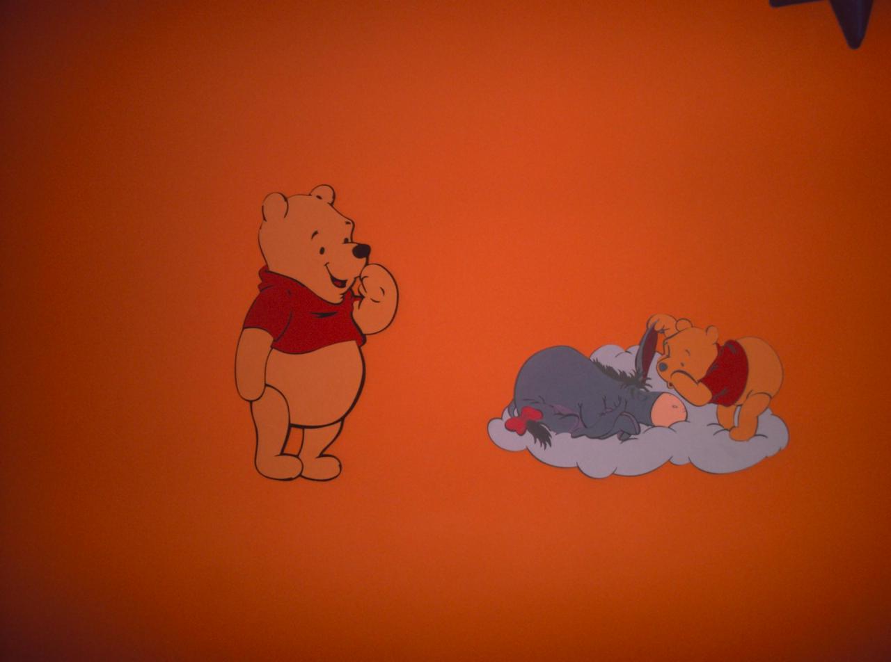 Ręcznie malowana na pomarańczowej ścianie postać Kubusia Puchatka w czerwonej koszulce i Kłapouchego śpiącego na szarej chmurce.