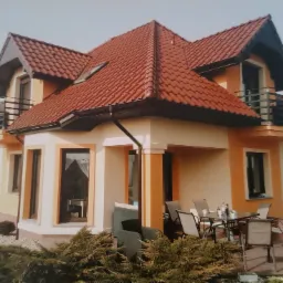 Dwukondygnacyjny dom jednorodzinny z balkonami, pomalowany na biało i pomarańczowo, z czerwoną dachówką, widok z poziomu gruntu na taras z meblami ogrodowymi i zadbanym otoczeniem.