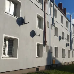 Szary blok mieszkalny z białymi obramowaniami okien i antenami satelitarnymi na elewacji, widok z poziomu chodnika w słoneczny dzień.