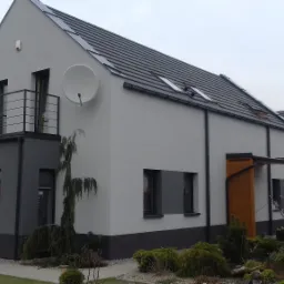 Dwukondygnacyjny dom jednorodzinny z balkonem, otynkowany na szaro z ciemnoszarą dolną częścią, widoczna antena satelitarna na elewacji, dach pokryty ciemną dachówką.