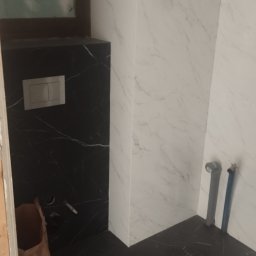 Usługi Remontowo-Budowlane Karol Gastoł - Wykończona łazienka z czarno-białymi płytkami imitującymi marmur. Widoczne przyłącza wodne i częściowo widoczny worek budowlany.