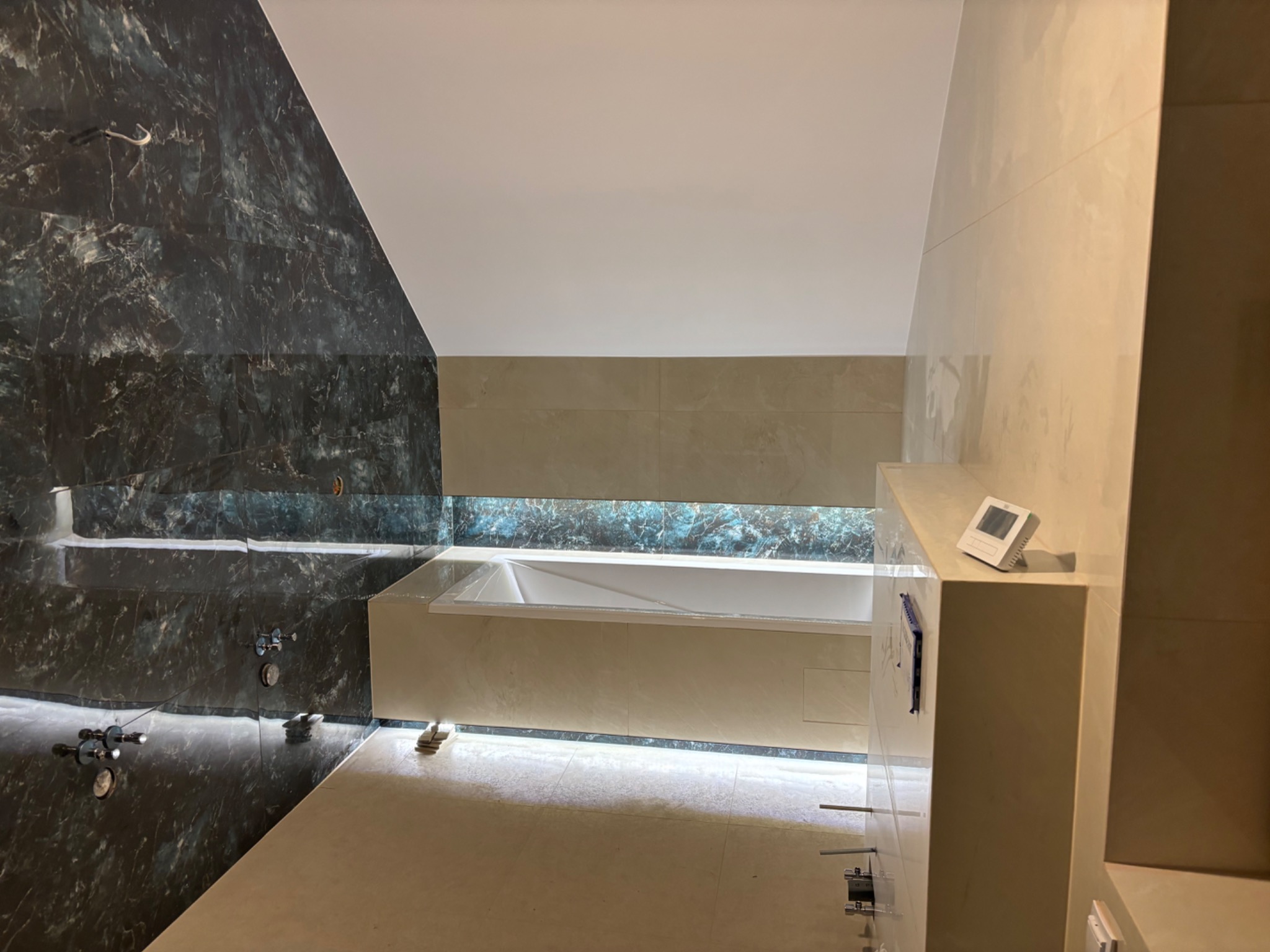 Wykończona łazienka z wanną, obłożona płytkami imitującymi marmur w odcieniach szarości i beżu, z oświetleniem LED. Widoczne detale armatury i panel sterowania.