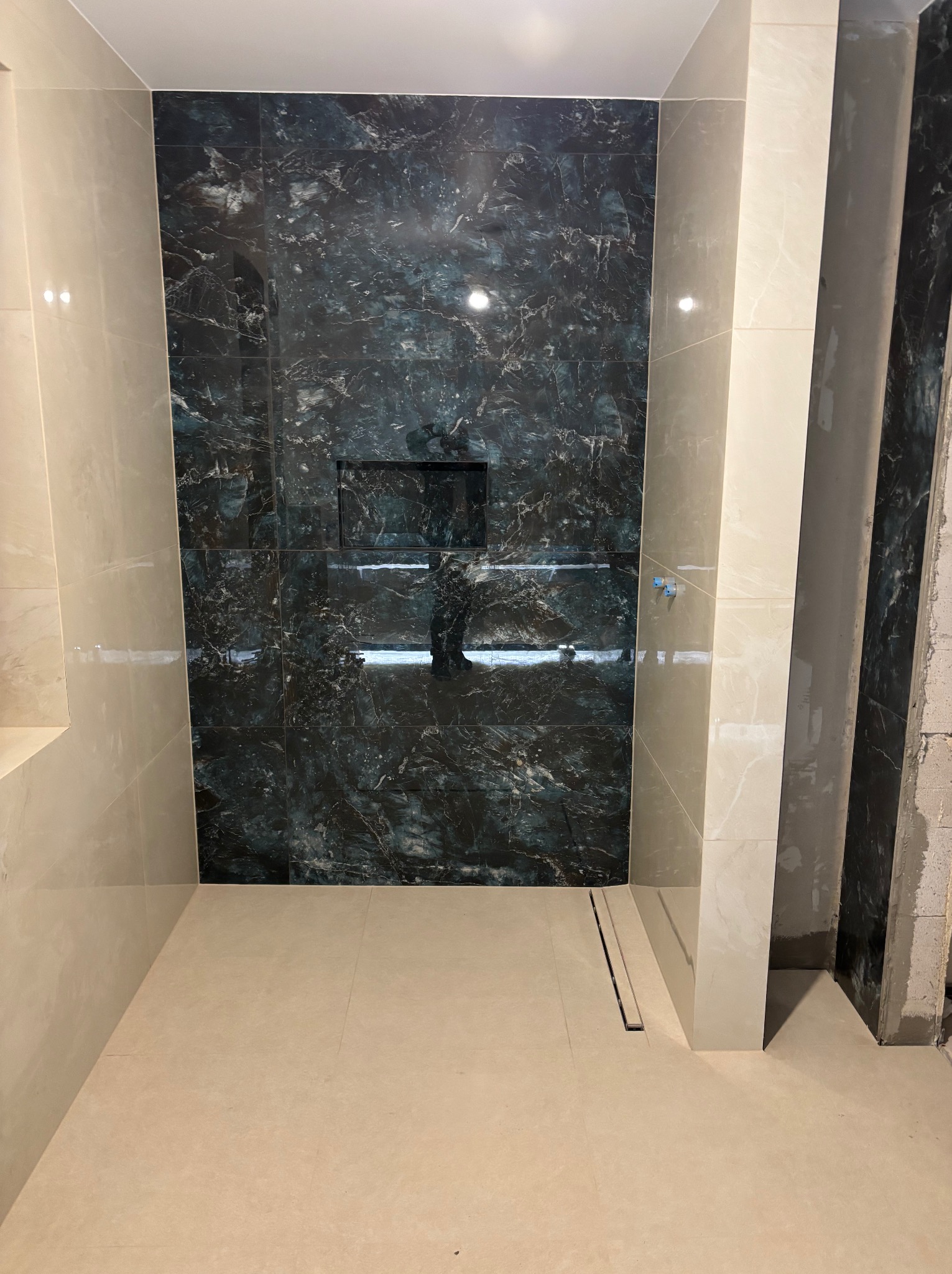 Wykończona kabina prysznicowa z ciemnymi płytkami imitującymi marmur i jasnymi płytkami po bokach. Widoczny odpływ liniowy w podłodze i wnęka w ścianie.