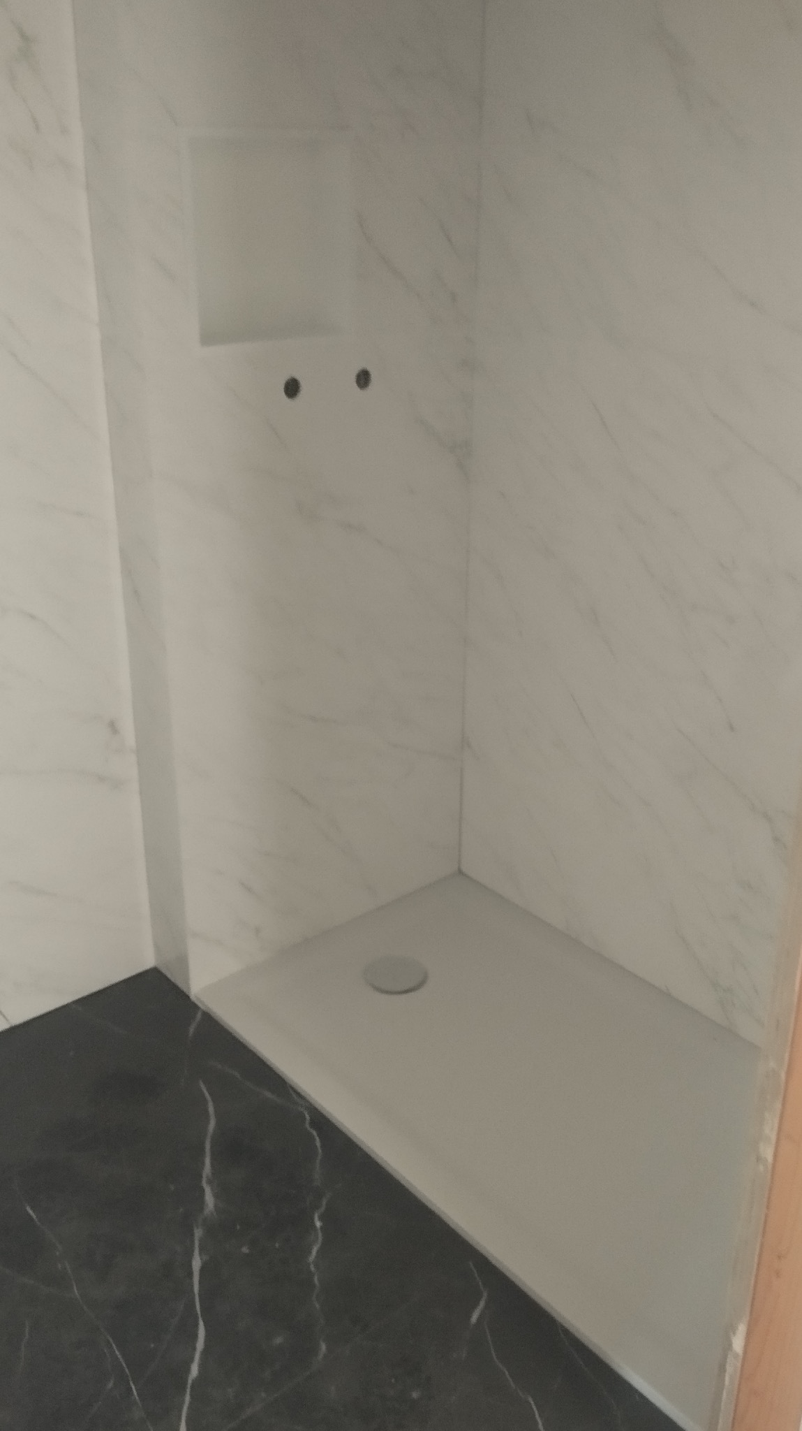 Wykończony brodzik z odpływem liniowym, obłożony marmurową płytą. Widoczna wnęka w ścianie. Podłoga wyłożona czarnym gresem z białym żyłkowaniem.