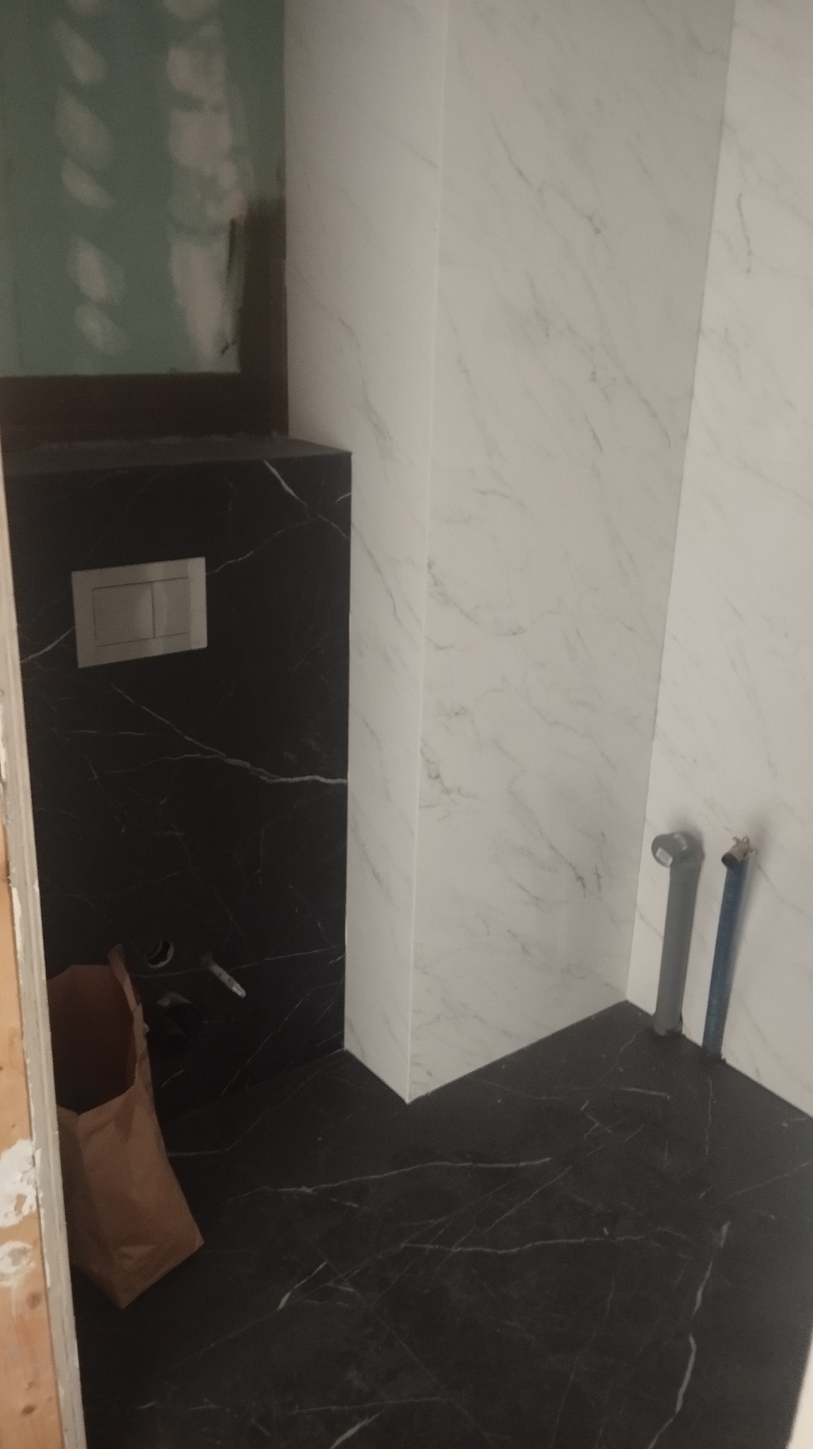 Wykończona łazienka z czarno-białymi płytkami imitującymi marmur. Widoczne przyłącza wodne i częściowo widoczny worek budowlany.