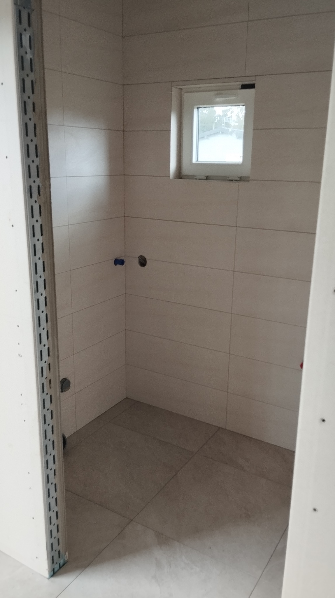 Wykończona łazienka z beżowymi płytkami na ścianach i podłodze. Widoczne otwory na armaturę i małe okno. Profil narożny na ścianie z płyty gipsowej.