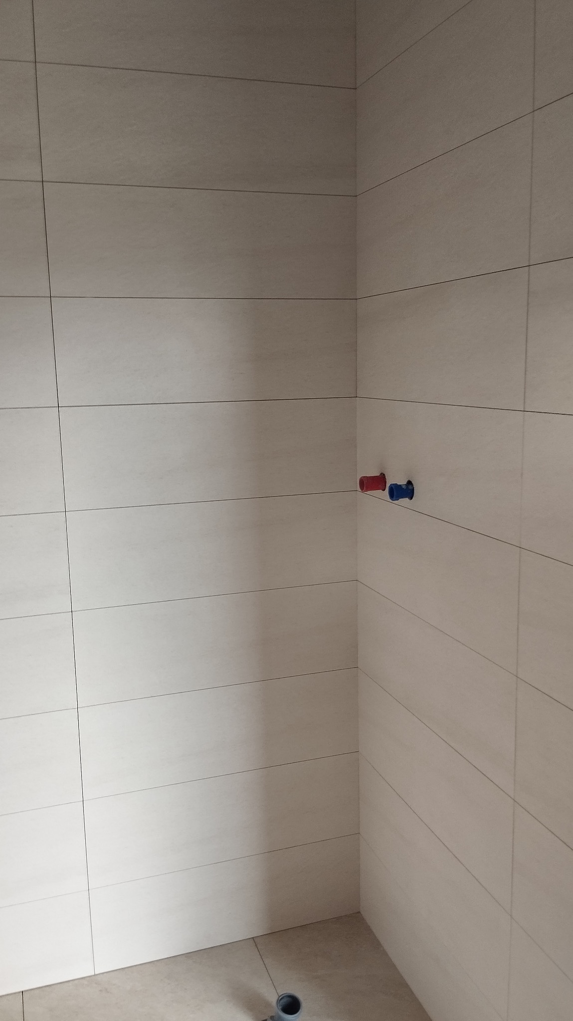 Wykończona łazienka z beżowymi, poziomymi płytkami. Widoczne przyłącza wodne (czerwone i niebieskie) oraz odpływ podłogowy. Minimalistyczny design.