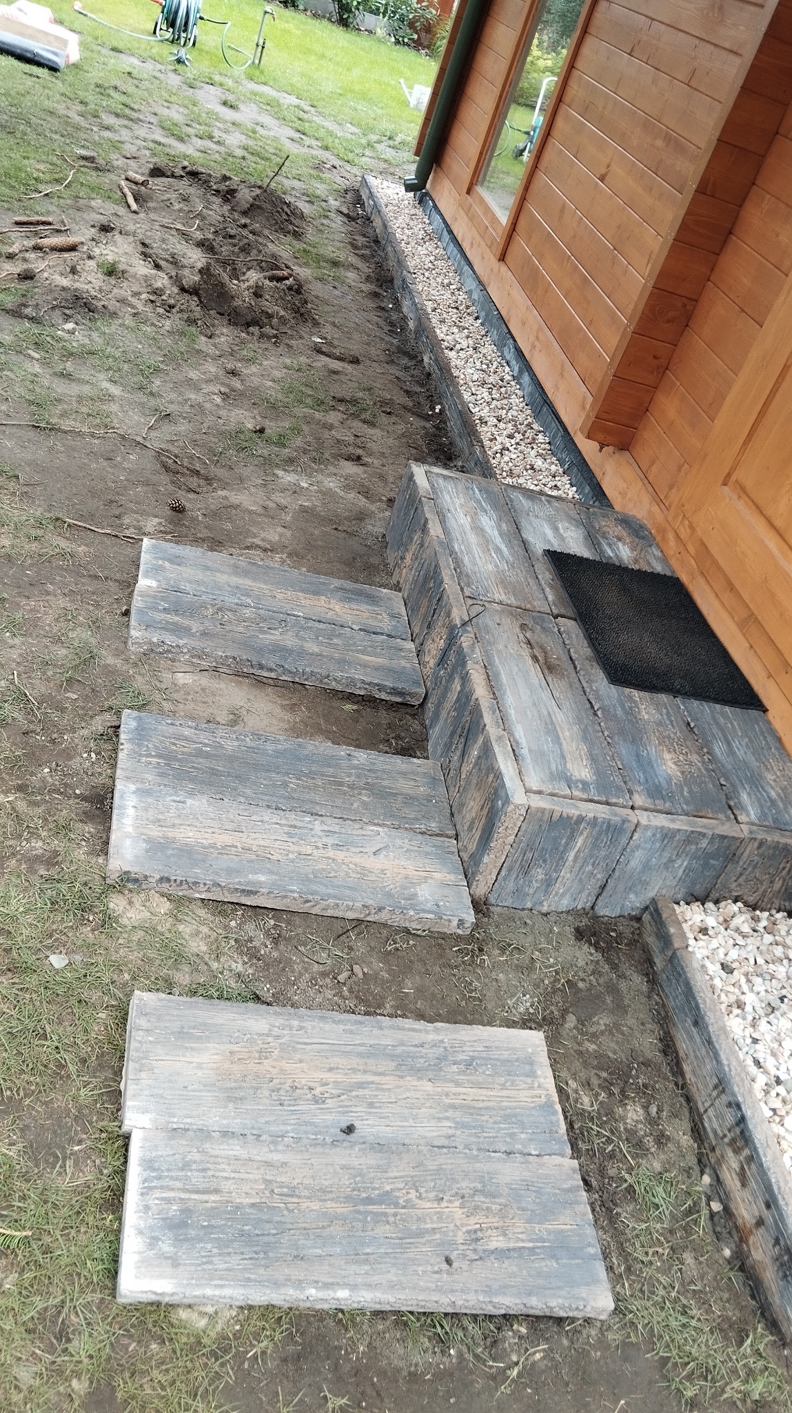 Ułożone stopnie z desek prowadzące do drewnianego domku, otoczone kamieniami i trawą. Widoczne detale wykończenia i naturalne materiały.