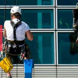 Pracownicy alpinistyczni myjący okna na wysokości w nowoczesnym biurowcu z użyciem lin, uprzęży, kasków i niebieskich wiader.