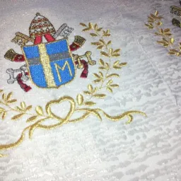 Biała tkanina z wyhaftowanym herbem papieskim otoczonym złotymi gałązkami, widoczne detale haftu i struktura materiału.