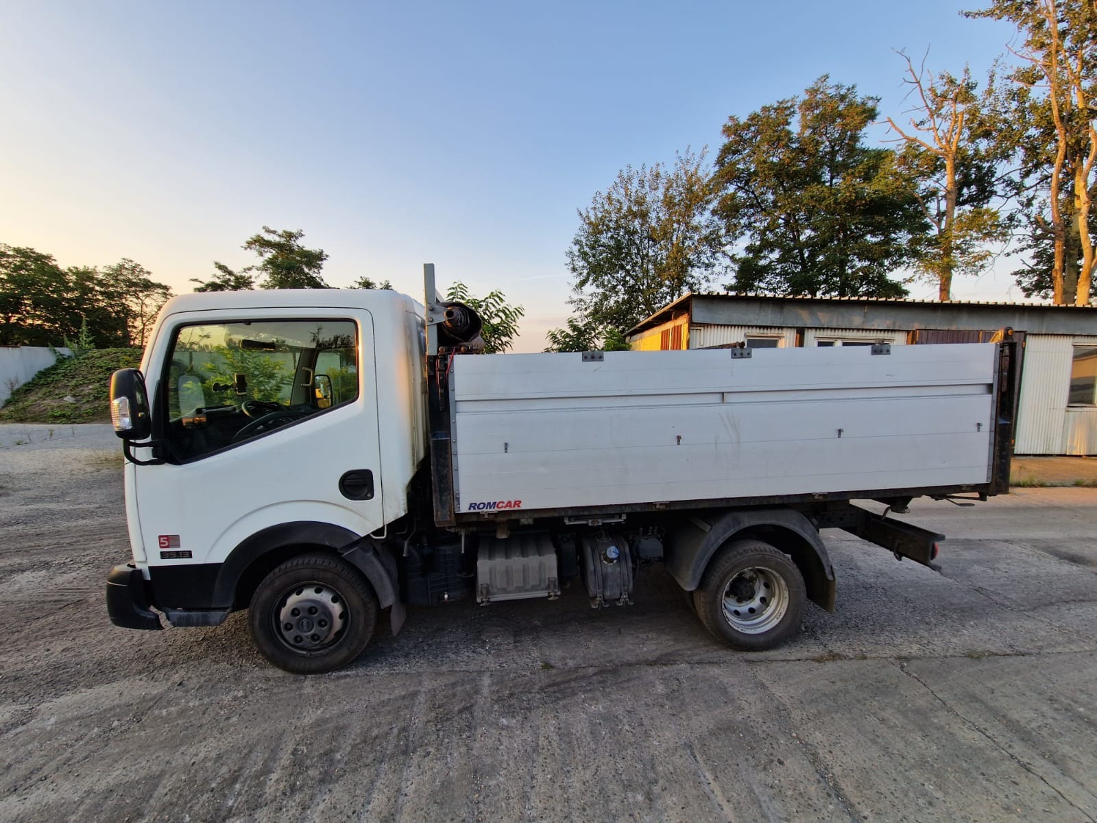 Biała ciężarówka z otwartą skrzynią ładunkową, zaparkowana na betonowym placu, w tle budynek i drzewa, widok z boku, oświetlenie popołudniowe.