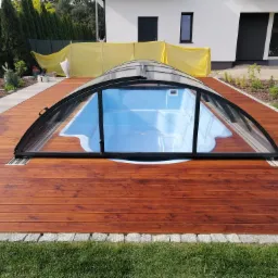 Zadaszony basen z błękitną wodą otoczony drewnianym tarasem z desek w odcieniach brązu, obrzeże z kostki granitowej, w tle trawnik i budynek z białymi ścianami.