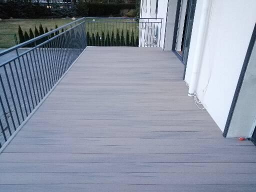 Szary taras z desek, z metalową balustradą, przylegający do białej ściany budynku z widocznymi drzwiami balkonowymi.