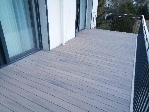 Nowoczesny taras z szarych desek kompozytowych z czarną metalową balustradą, widok z góry na tle białej ściany budynku i zieleni.