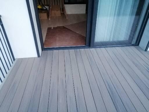 Szare deski tarasowe z widocznym fragmentem balustrady i otwartym oknem przesuwnym, wnętrze domu w tle.