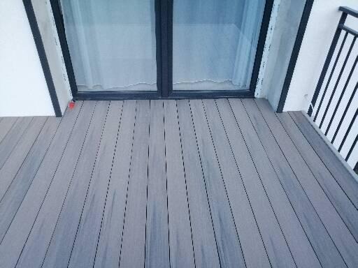 Szare deski tarasowe ułożone na balkonie, widoczne drzwi balkonowe i fragment balustrady.