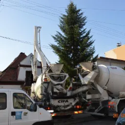 Dwie betoniarki CIFA na budowie, widoczny fragment domu z czerwoną dachówką i zielone drzewo iglaste w tle.