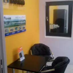 Ekspozycja okna w ciemnej ramie w salonie sprzedaży Dako, z widocznym logo i materiałami reklamowymi na żółtej ścianie.