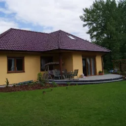 Parterowy dom z żółtą elewacją i bordowym dachem, taras z meblami ogrodowymi, zadbany trawnik, widok na ogród i drzewa. Ujęcie w słoneczny dzień.