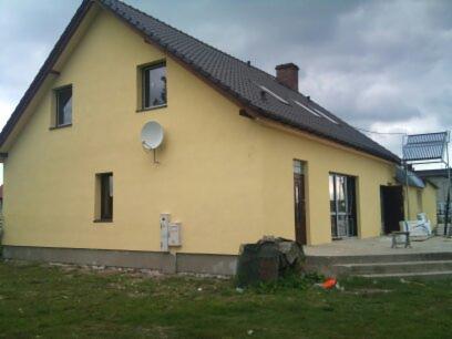 Dom jednorodzinny po nałożeniu żółtego tynku zewnętrznego, widoczna antena satelitarna, okna i drzwi z ramami w kolorze ciemnego drewna, panele solarne na dachu budynku gospodarczego w tle.