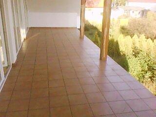 Wyłożony brązowymi płytkami taras z drewnianymi słupami podtrzymującymi zadaszenie, widok na zieleń w tle.