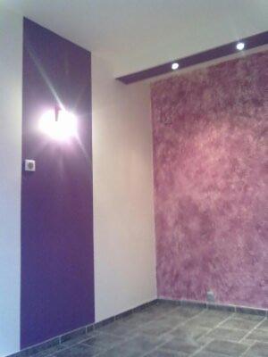 Pokój po odświeżeniu: ściana w odcieniach fioletu z dekoracyjną strukturą, pionowy pas w kolorze fioletowym obok białej ściany, szara podłoga z płytek, oświetlenie sufitowe.