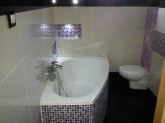 Biała wanna narożna z armaturą, toaleta i fragment ściany z mozaiką w odcieniach fioletu i bieli, tworzące wykończenie łazienki.