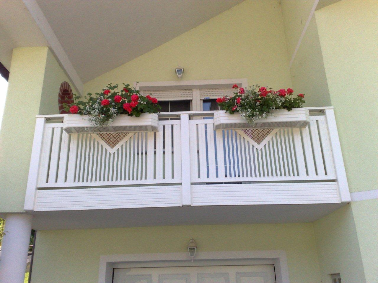 Biała, drewniana balustrada balkonu z dwoma skrzynkami kwiatowymi z czerwonymi i białymi kwiatami, widok z dołu na jasnym tle budynku.