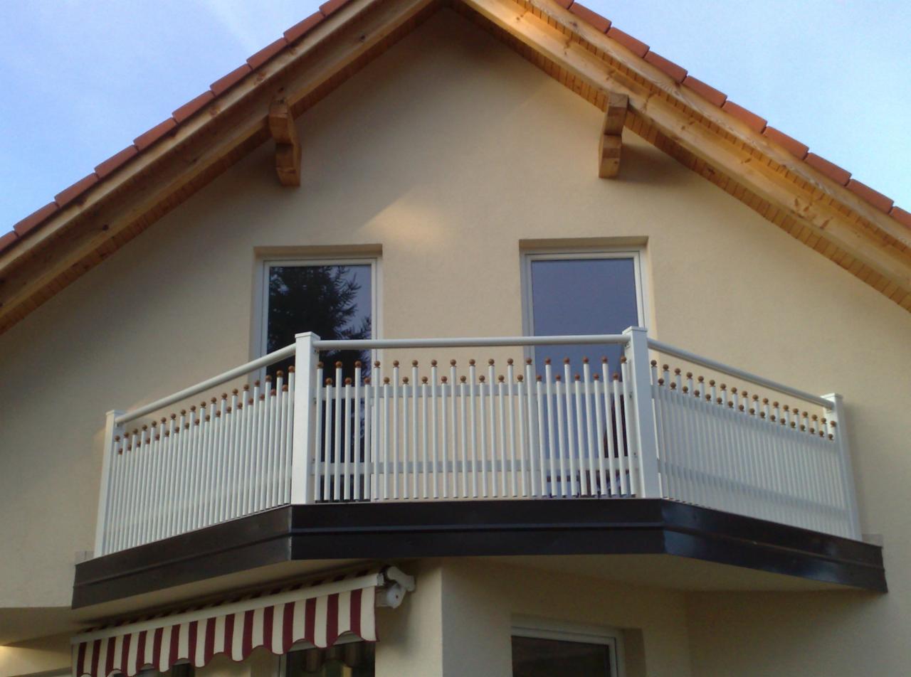 Biała, metalowa balustrada balkonowa z ozdobnymi kulkami na tle beżowej elewacji domu z dwoma oknami i czerwonym dachem.